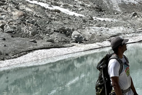 Man outdoors wearing a hat and backpack. The landscape is blue water surrounded by melted snow on a mountain.